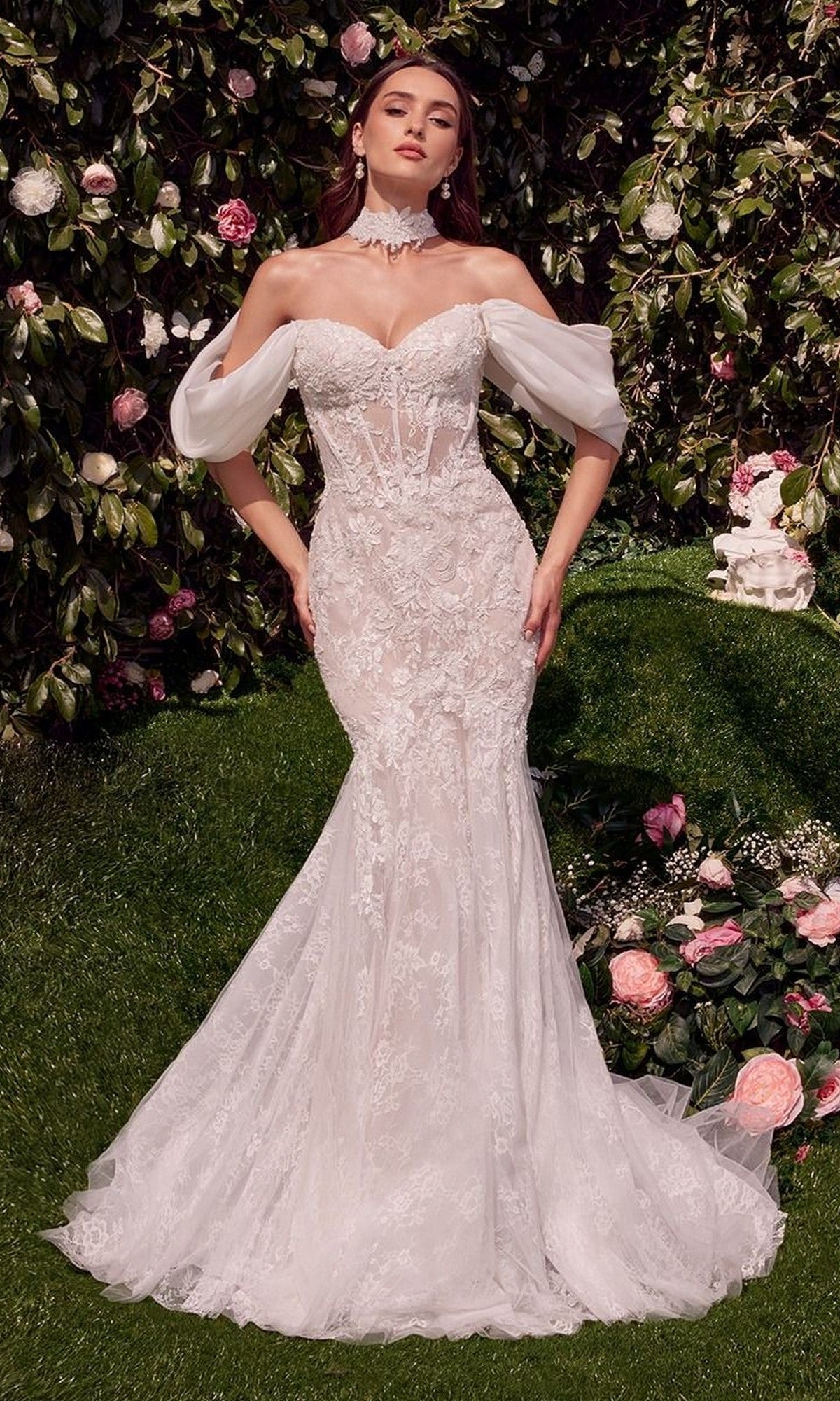 A woman in the Andrea and Leo WL031 long wedding dress in off-white, featuring a strapless lace mermaid design with sheer sleeves and a removable organza shawl, stands in a blooming garden.